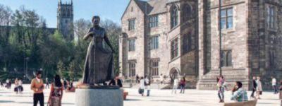 A statue of a woman in a long dress stands in a paved square in front of a large stone building with many windows. People walk around the square under a clear blue sky.